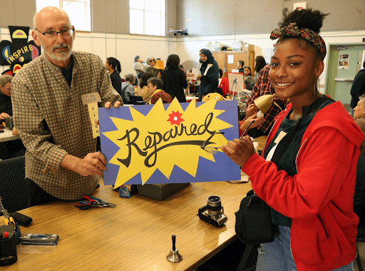 Two people at a Repair Cafe holding a sign that says "Repaired". On the table between them is a bell, a camera and some tools.