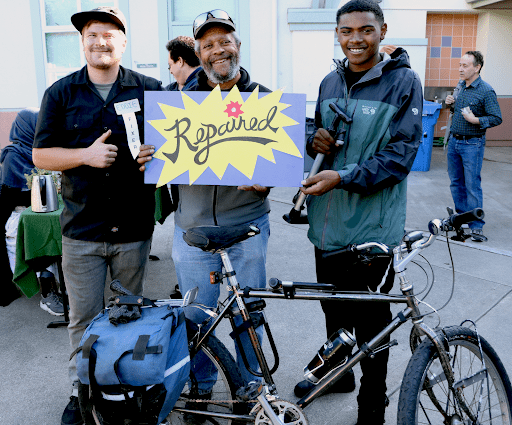 Three men in the foreground with a bicycle in front of them. One is giving a thumbs up, one is holding a sign that says "Repaired", and the third is holding a bicycle pump