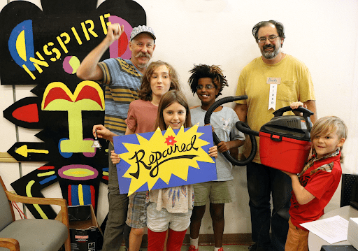 Two men and four children feature in the photo, with an art work behind them that says "INSPIRED". One man has his fist in the air, one child is holding a bell, two children are holding a sign that says "Repaired", and a fourth is holding up a vacuum cleaner.