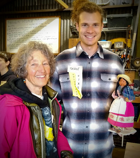 Two people photographed in a repair cafe. Both smiling to camera one has a name badge which says "Sean - Volunteer Fixer", and he is holding a doll.